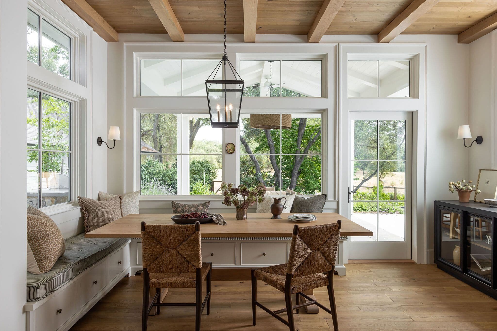 A photo of a large window in a dining area with a wooden kitchen table, chairs, and banquette for seating